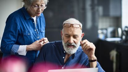 An older man looks at a laptop on the kitchen table while his wife looks over his shoulder.