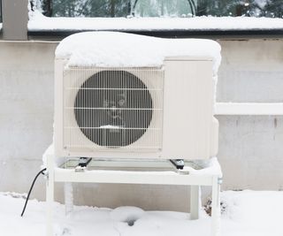 A heat pump unit covered in snow in front of a light wall of a home