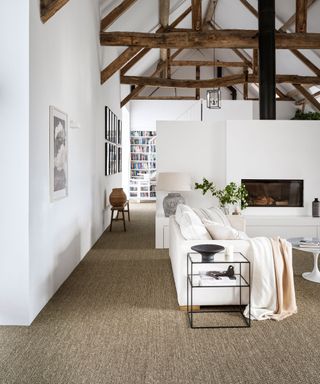 Large open plan white living room, barn conversion, vaulted ceiling with dark wood beams on show, seating area around fireplace, textured, natural carpet, view onto library area with white staircase