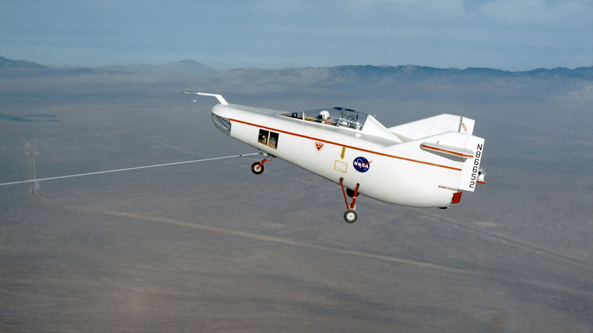 a small white aircraft with a pilot aboard is towed in flight by line extending to the dry lake bed below.
