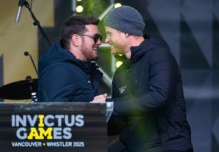 Michael Buble and Prince Harry hugging and wearing black winter coats on stage in front of an Invictus games podium