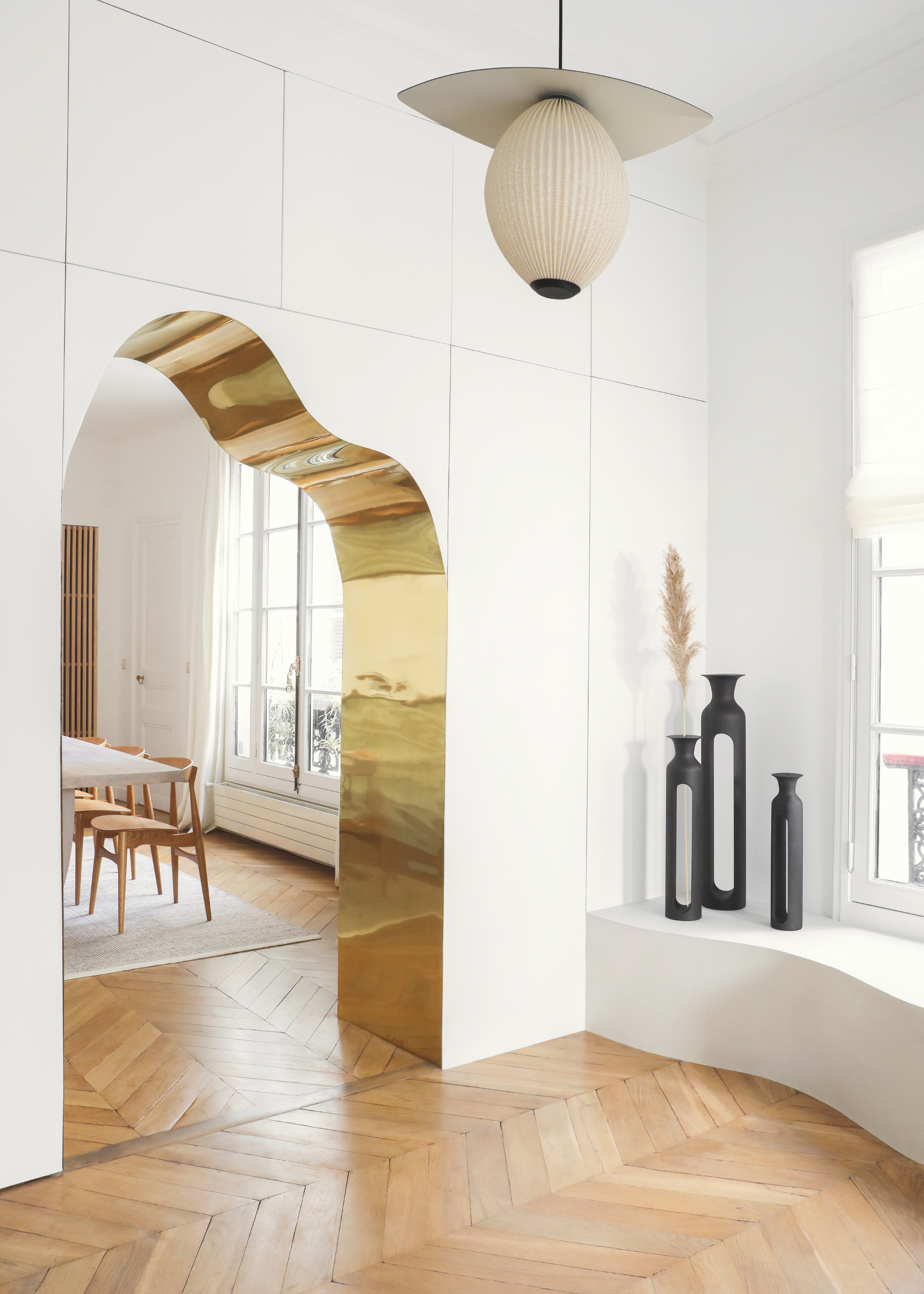 a wavy golden archway separating a fully white living and dining space with pale herringbone wood flooring and black statement ornaments in the corner and a statement ceiling light