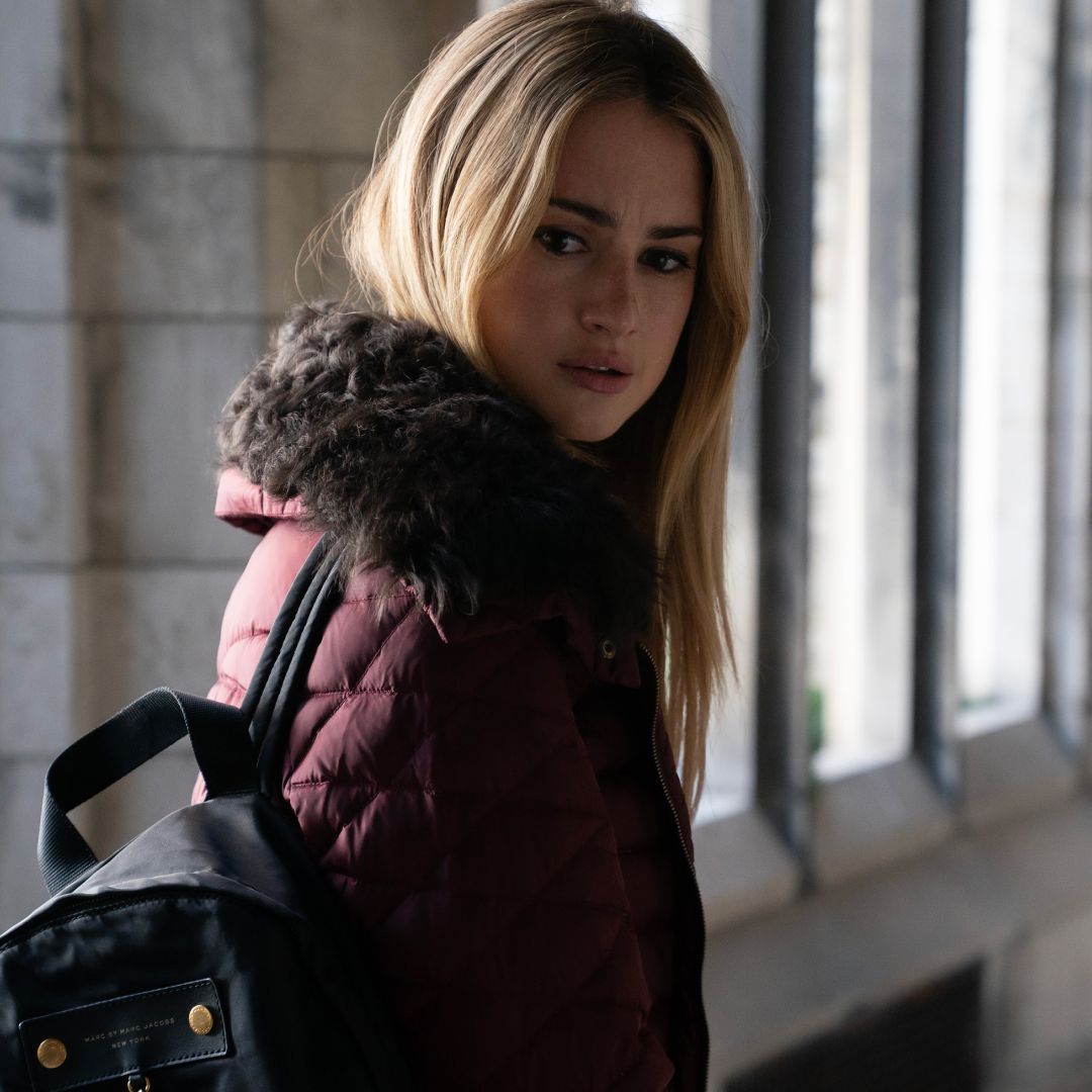 grace van patten as lucy ewaring a fur lined coat walking down a college hallway looking over her shoulder in tell me lies season 3