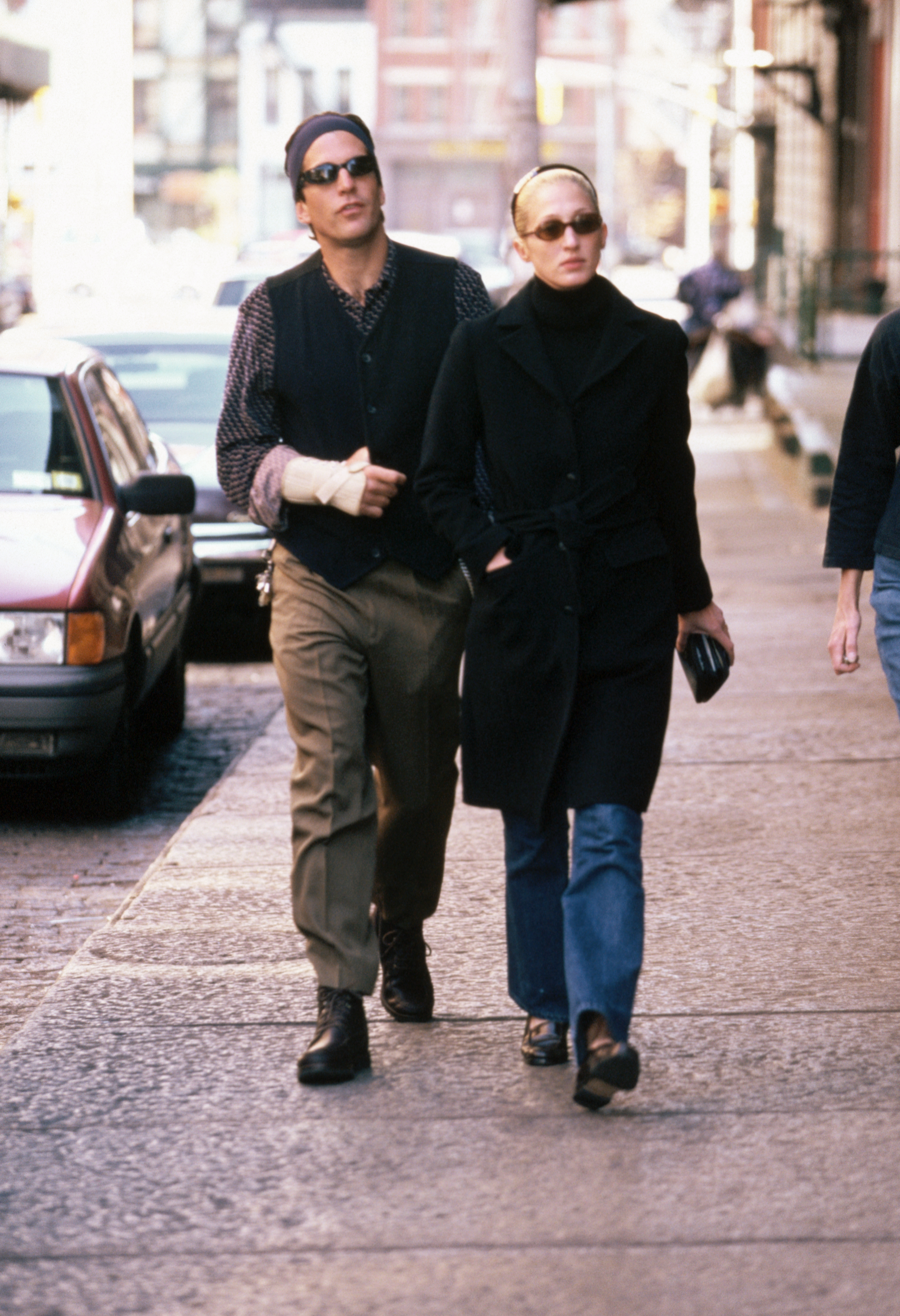 John F. Kennedy Jr. walks along a New York City street with wife Carolyn Bessette, who wears blue jeans, a black coat and black turtleneck and wears a wide headband in her blonde hair