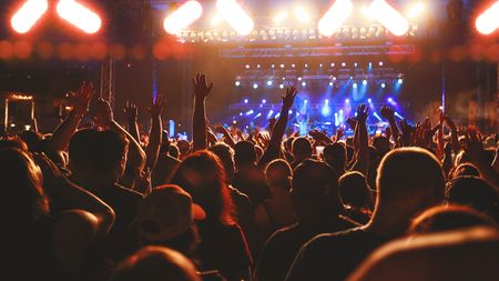 Concert crowd cheering, concert audience arms raised. Live entertainment concept of music festival crowd cheering for live music performance, rock music concert event, or enthusiast fans enjoying nightlife. Rear view concert crow, audience with concert lights and stage background. Part of a series.
