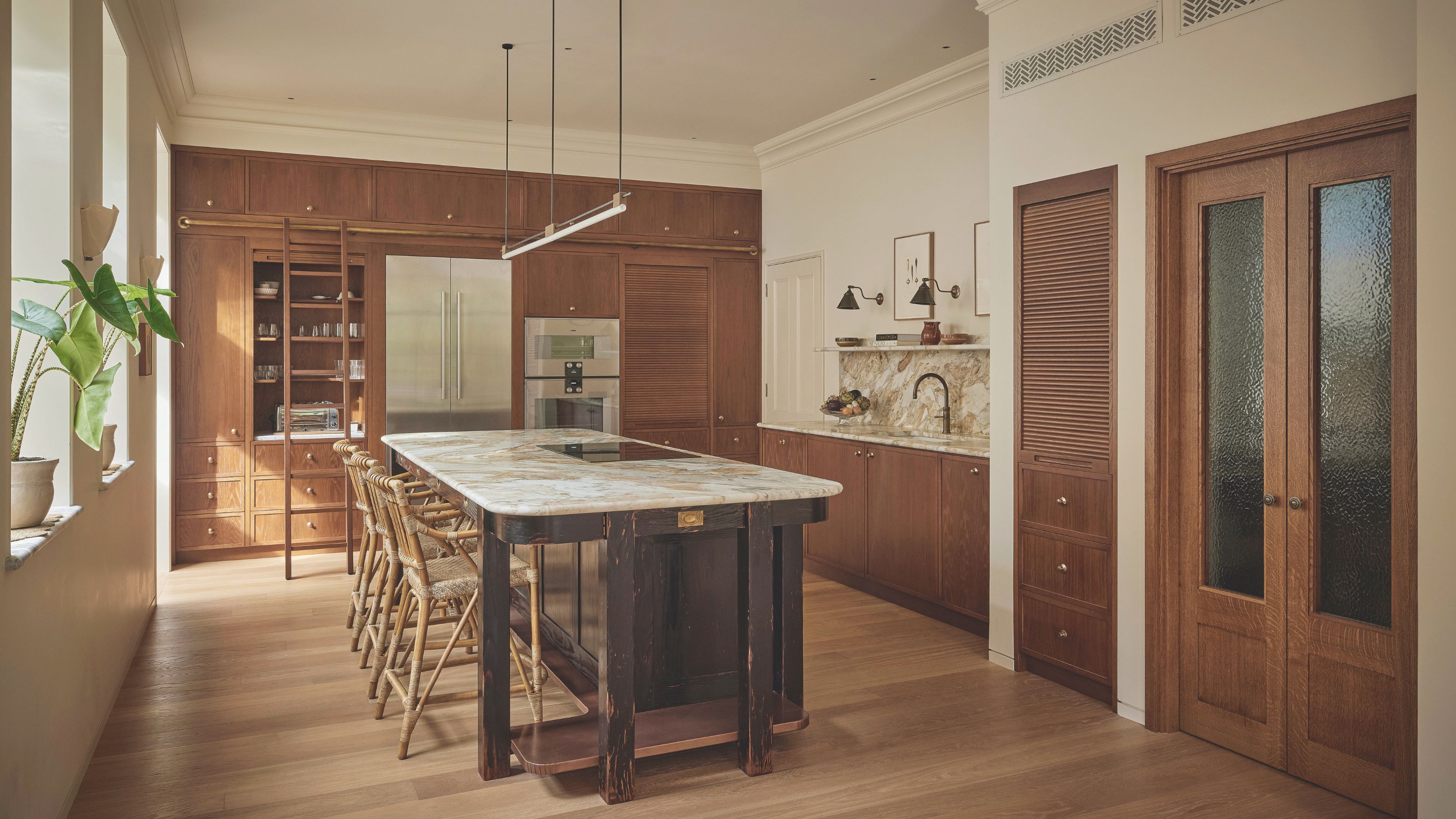 A light filled kitchen with cream walls and wooden cabinetry. A darker wood kitchen island sits in the center with a white marble countertop