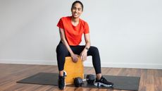 Woman smiling at viewer while sitting on wooden plyo box with a dumbbell between her feet
