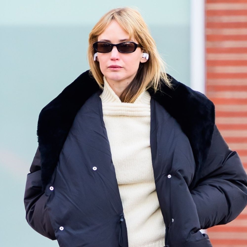 Jennifer Lawrence walks down the street in New York wearing a puffer jacket with a butter yellow jumper and a pink scarf tied around her waist.