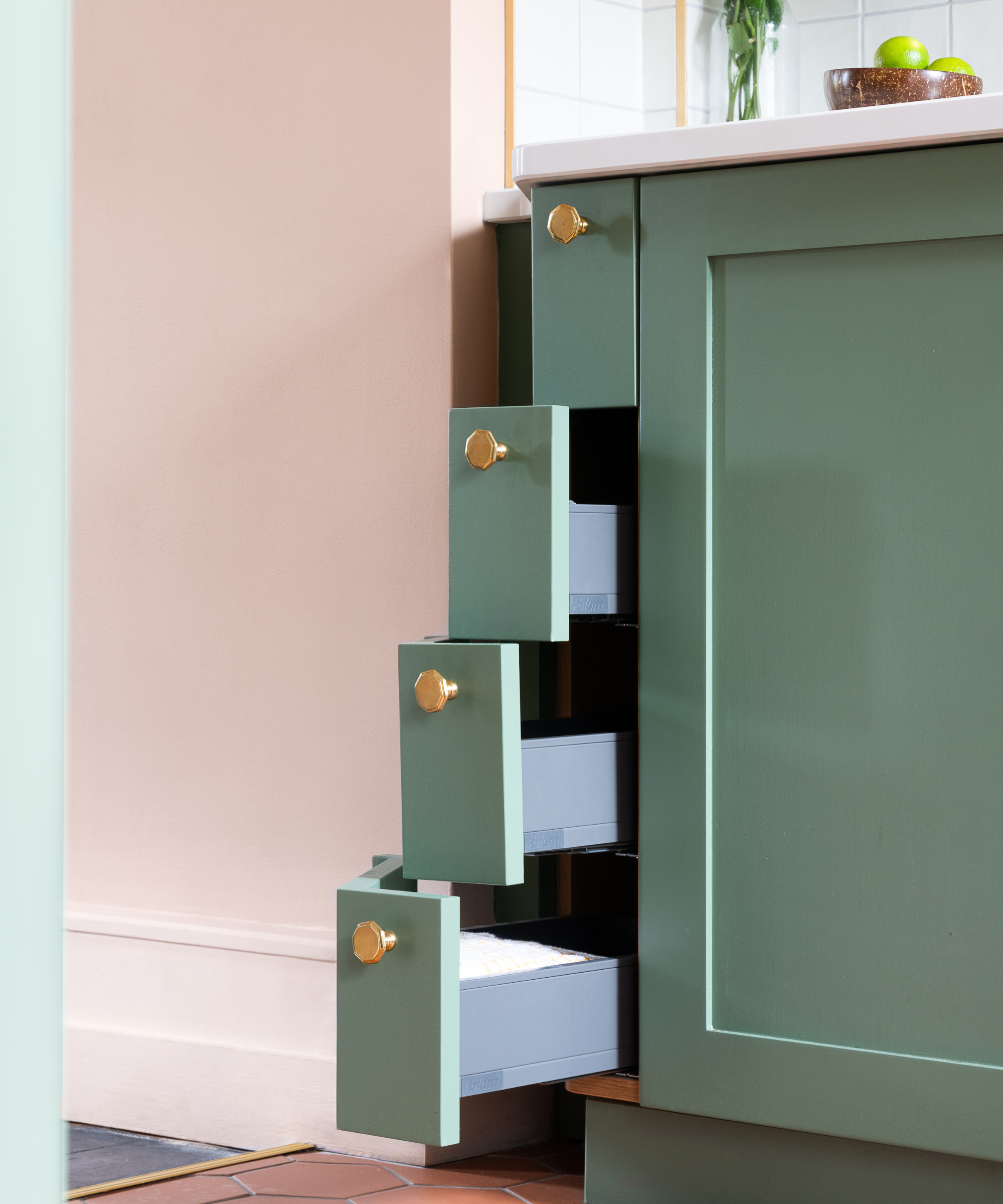 A set of slim, hidden drawers opens from the side of a green kitchen island, each fitted with gold knobs.