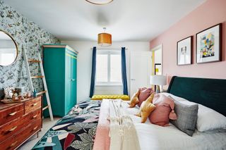 Bedroom with one pink wall and one patterned wallpapered wall, green velvet bed, bedding with yellow and pink cushions, and green painted wardrobe