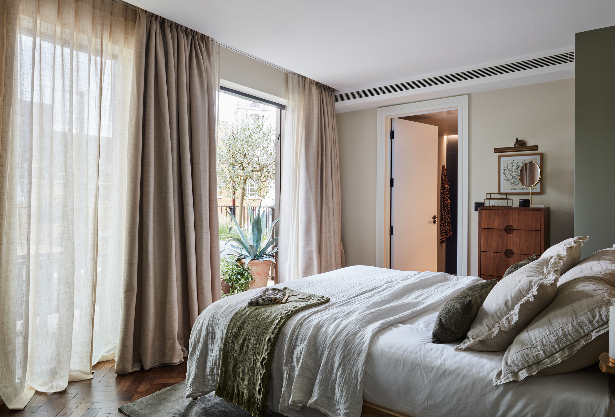 A serene bedroom with white linen bedding, a green throw blanket, floor-to-ceiling beige drapes, dark wood herringbone floors, and open doors leading to a potted plant-lined balcony.