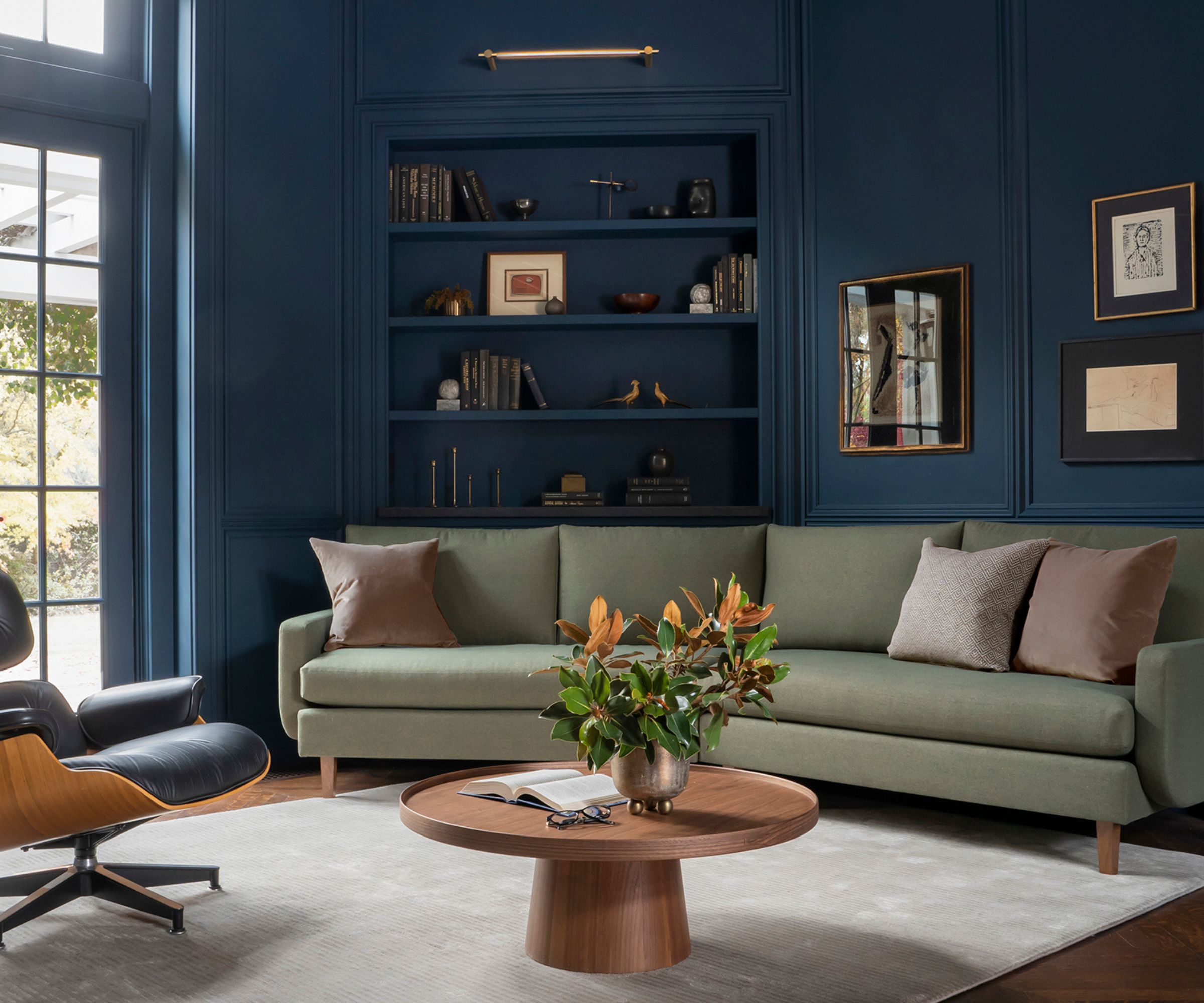 Home office with dark blue walls and shelves, green sofa, wood round coffee table, eames chair and paintings