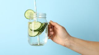 Image of someone holding a glass of cucumber water against a blue backdrop