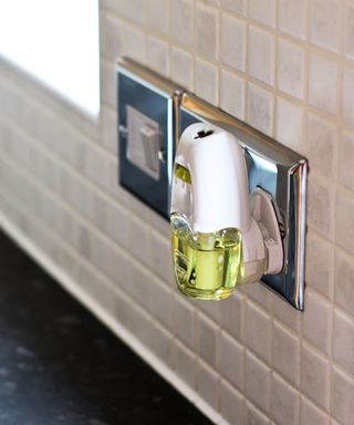 A close up of a white plug-in air freshener on a silver base with white tiles on the wall