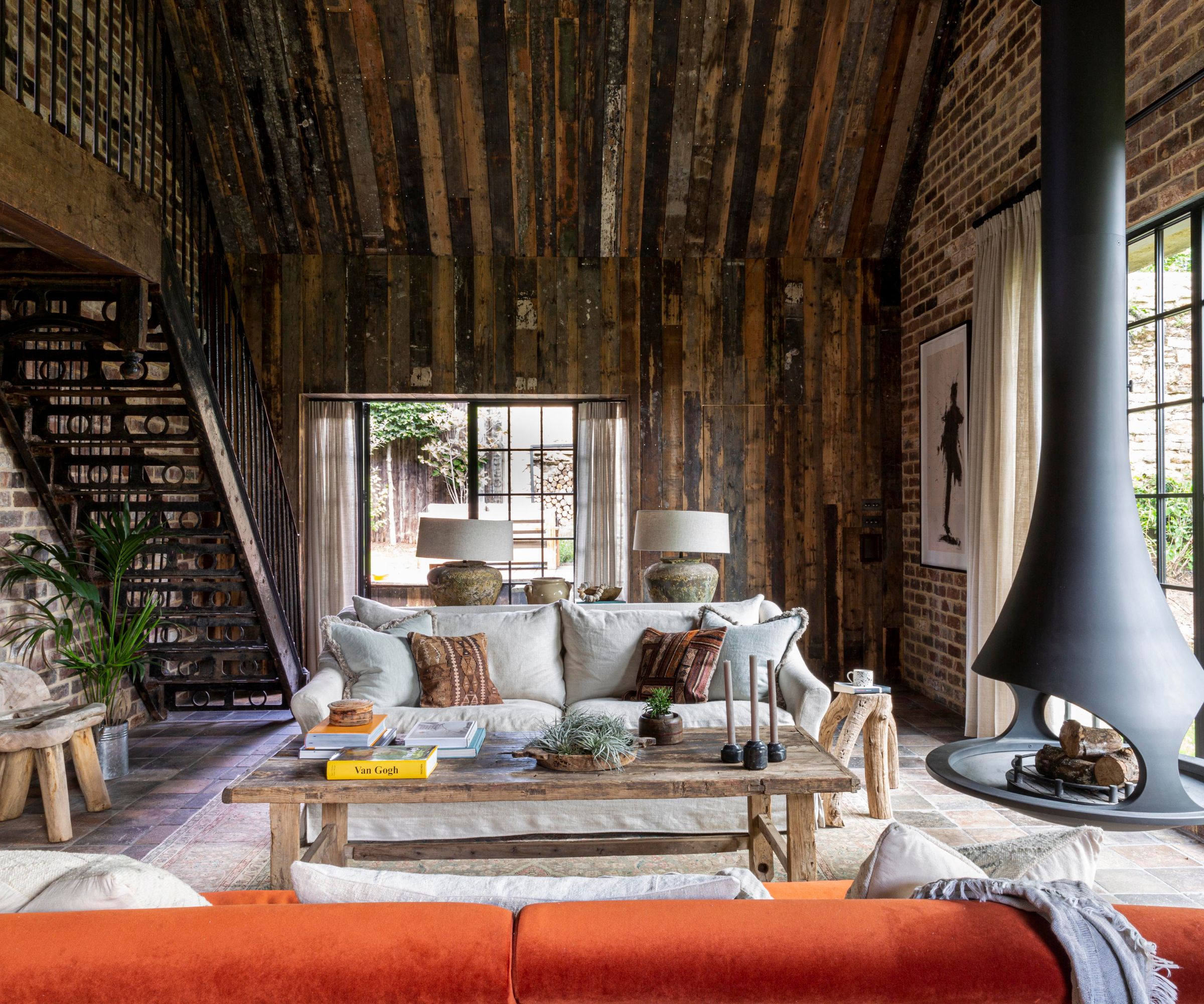 large country barn style living room with rustic wood paneling, exposed brick walls, and a large contemporary log fire hanging from the ceiling with a statement orange couch
