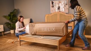 The image shows two women sliding the Helix Twilight mattress out of a cardboard box and placing onto a wooden bed frame