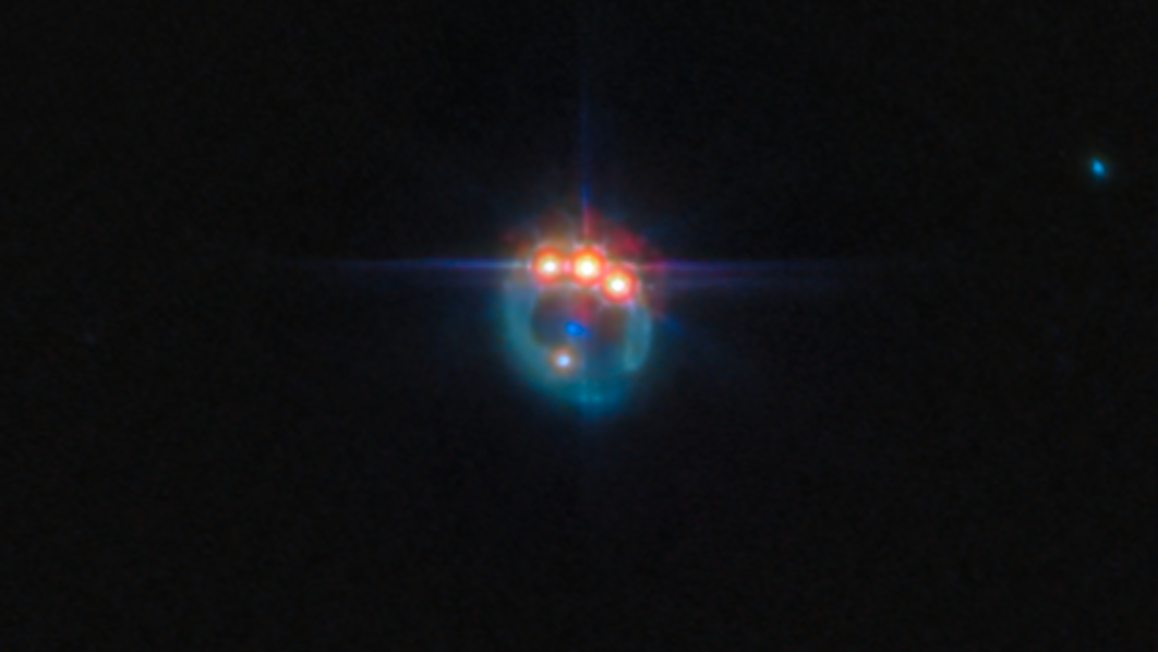 A blurry teal colored gas ring in black space features three bright shining orange orbs at its crown. A blue light dimly shines from the ring&#039;s center, and a paler small dot also shines on the right side of the image.