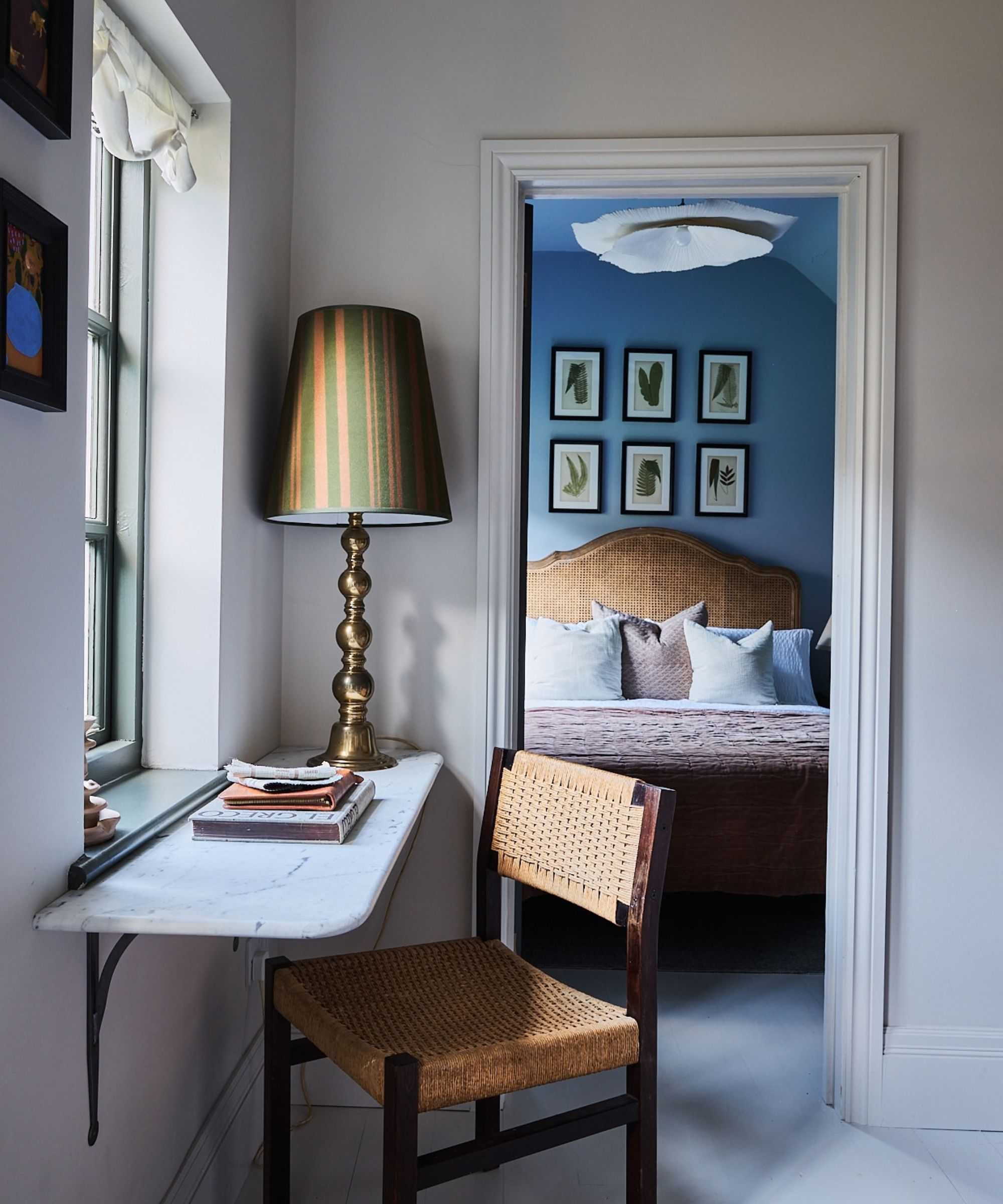 Attic study with white walls, a marble desk, a rattan chair, and a large table lamp with a green and yellow shade, looking into a blue bedroom with a rattan headboard, and six prints on the wall.