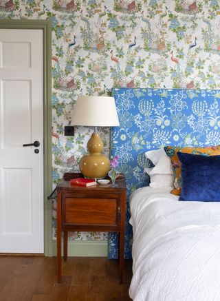 vibrant patterned green bird print wallpaper and bright blue patterned headboard in bedroom with blue velvet cushion and bedside table and lamp