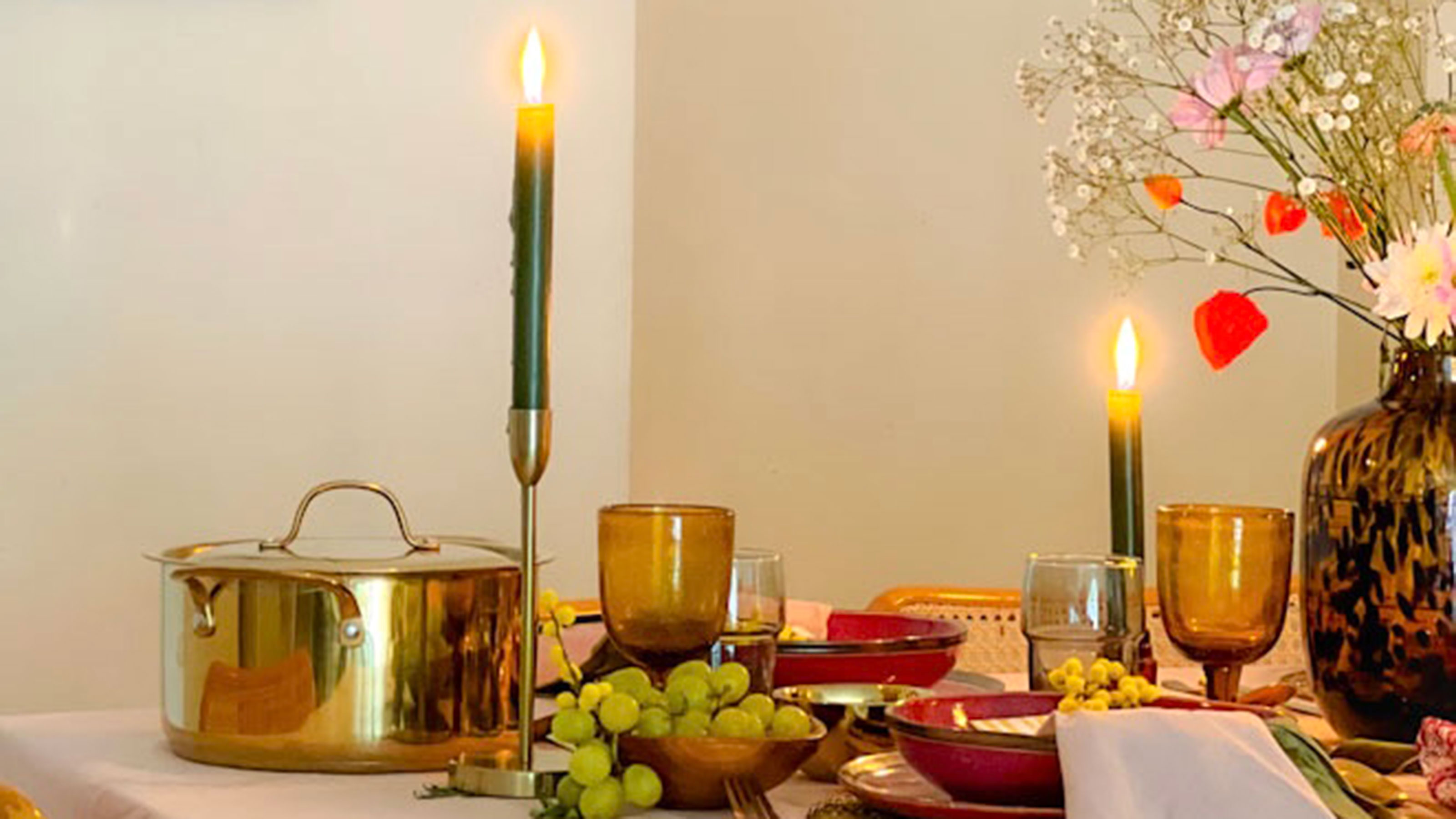 Table with tablecloth, metallic pot, golden candlesticks with lit pillar candles, amber wine glasses and tortoiseshell vase with fresh flowers