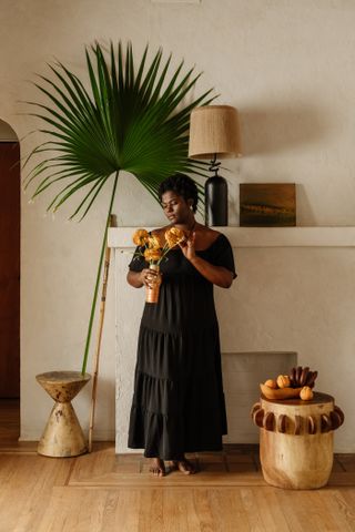 A woman in a black maxi dress holds a bunch of orange flowers while standing between different wooden stools and right underneath a glass and rattan lamp which sits on a shelf.