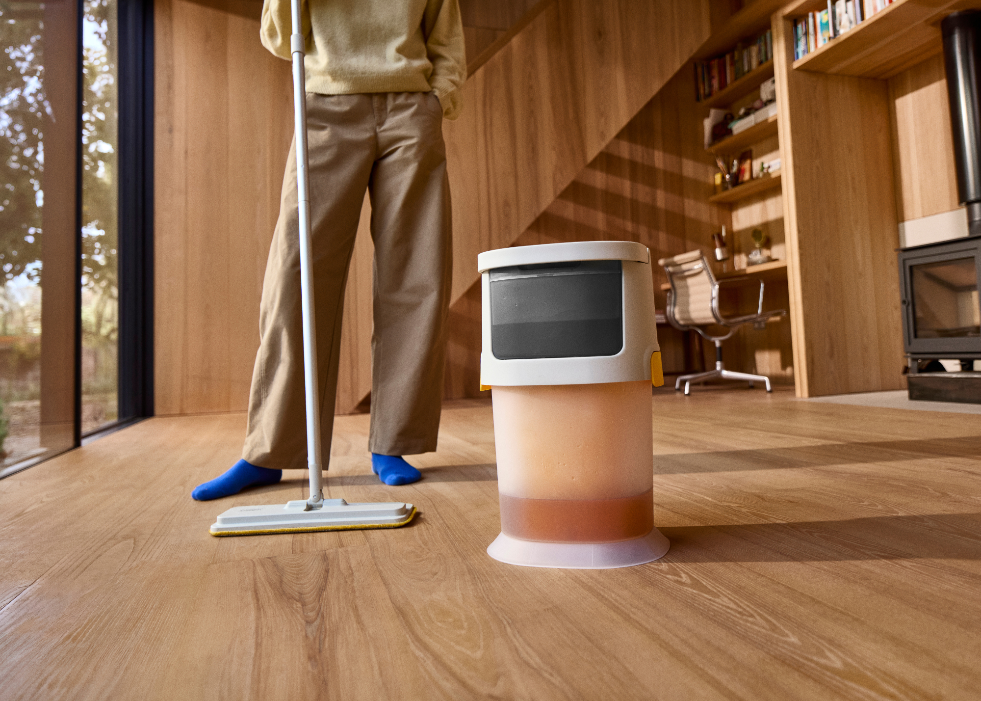 A Joseph Joseph UltraClean mop on a wood living room floor beside a person wearing khaki pants and blue socks