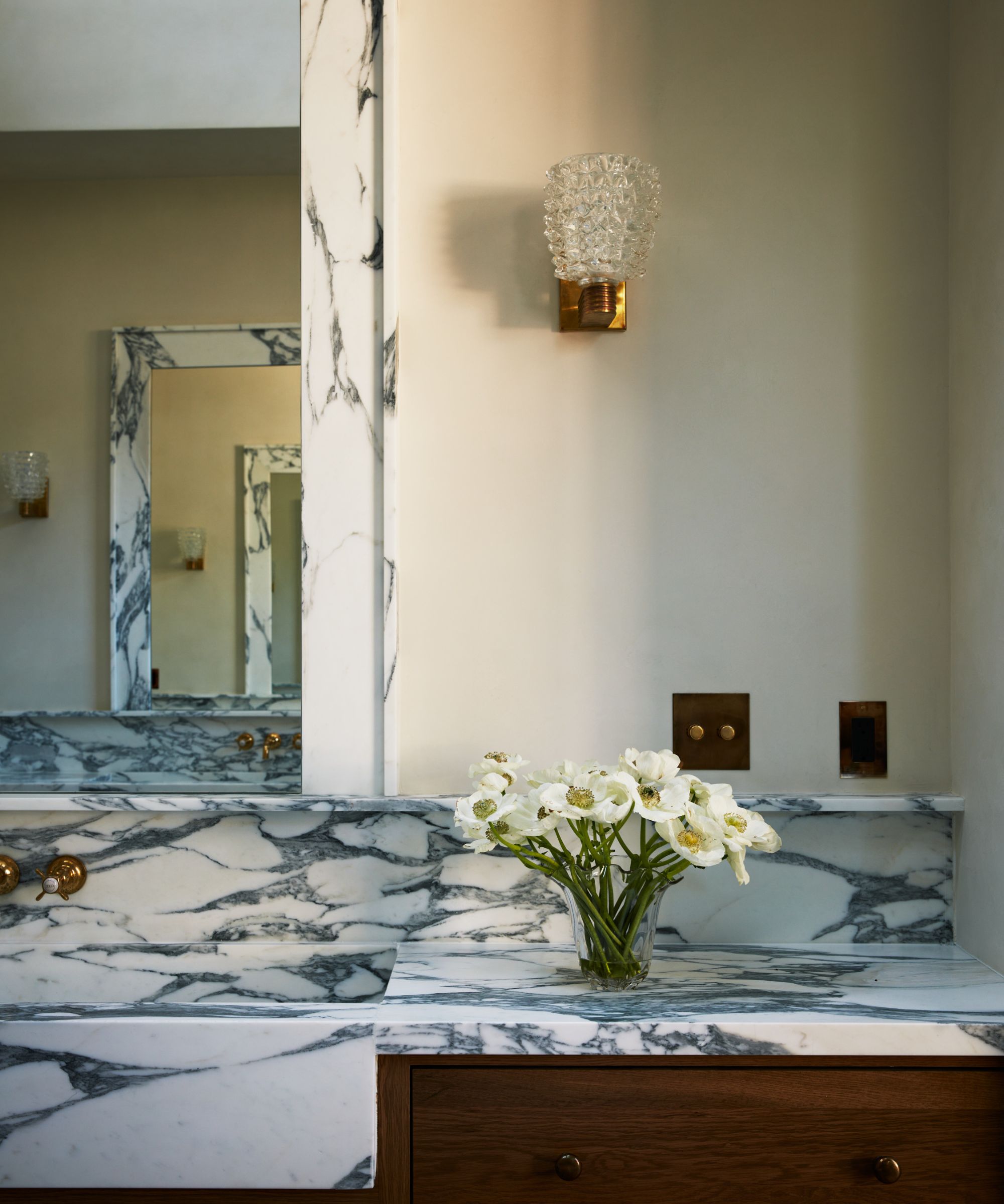 Close up of marble vanity in bathroom, vase of flowers placed on top, large mirror to the left, wooden cabinetry below