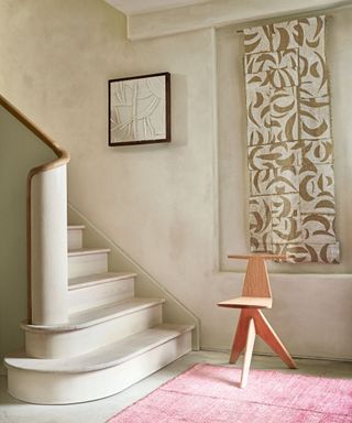 Calming hallway with textured, neutral painted walls and staircase, hanging patterned tapestry, artwork, wooden chair, pink rug