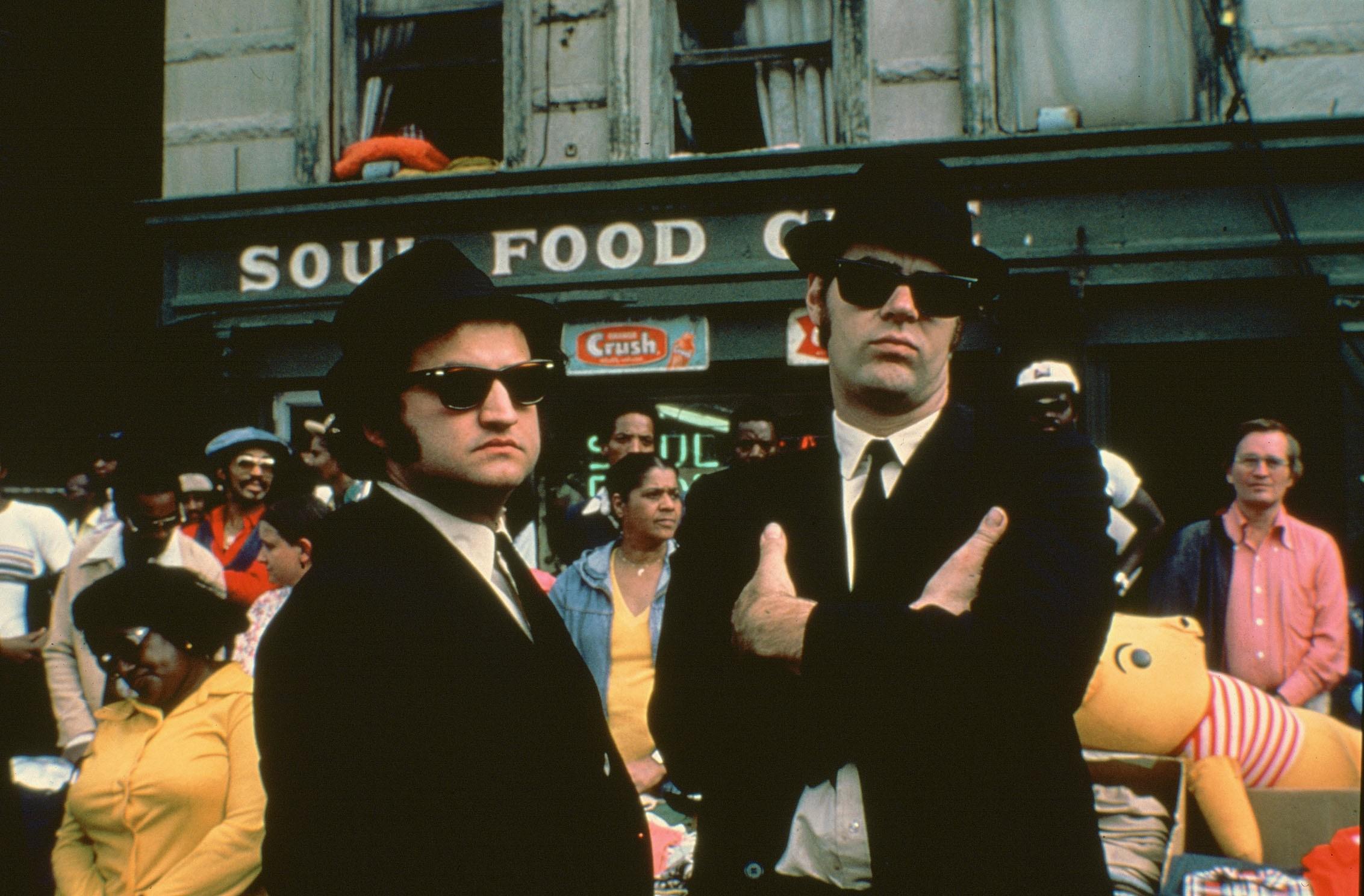 Two men in black suits and hots and sunglasses stand in front of a busy market