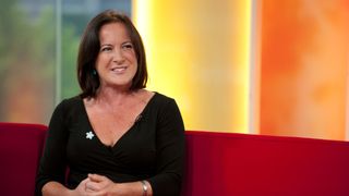 Cathy Murphy in a black dress, smiling and sitting on a red sofa on a talk show.