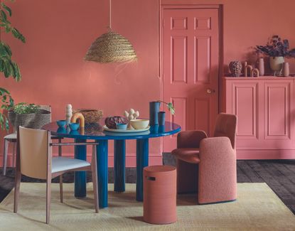 Dining area with red walls, dark floor and green rug, blue table and coloured chairs.