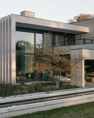 The front of a double storey square shaped architecturally design house with, large glass walls, a balcony, an outdoor entertainment area and plants and trees in front of it.
