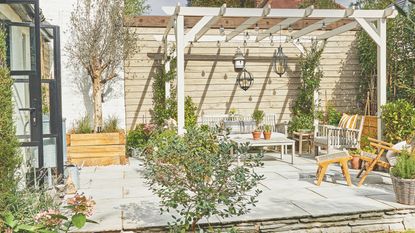 Large patio area covered in a wood pergola with garden furniture and plants around it