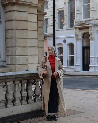 Mira wears a trench coat, red jumper, black maxi skirt and loafers