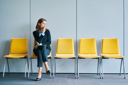A woman in business clothes sits waiting in a room with yellow chairs.