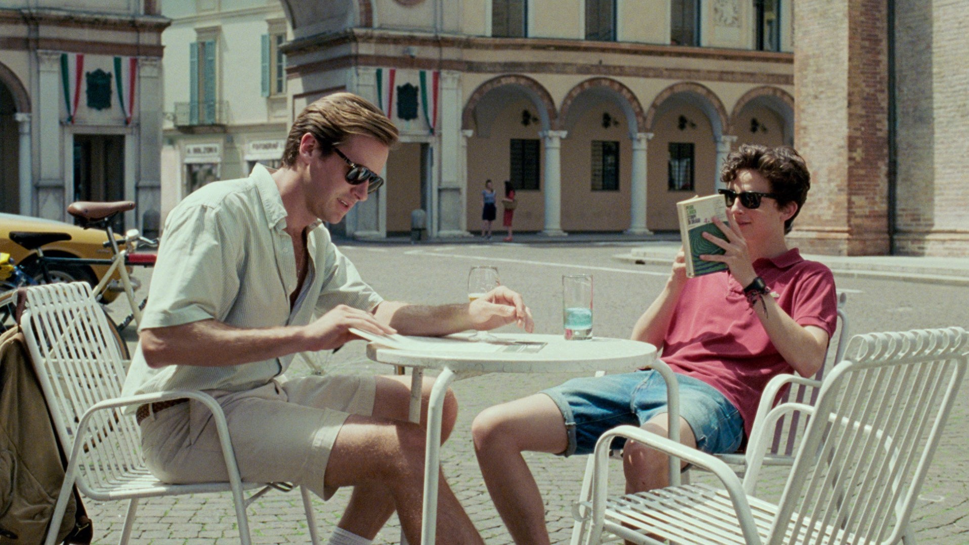 A still from the movie Call Me By Your Name, of main characters Elio and Oliver sat in a town square on white chairs.