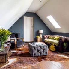 A converted loft with white angled ceiling, dark blue walls, wooden floors, a brown leather sofa, and animal skin rug