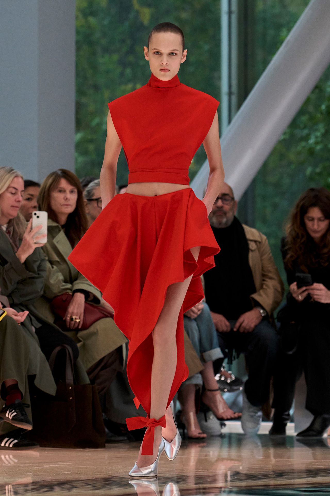 An Ala&iuml;a model wearing a bright red cap sleeve turtleneck top styled with matching red shorts that drape down to the ankles and a pair of silver pointed mules.