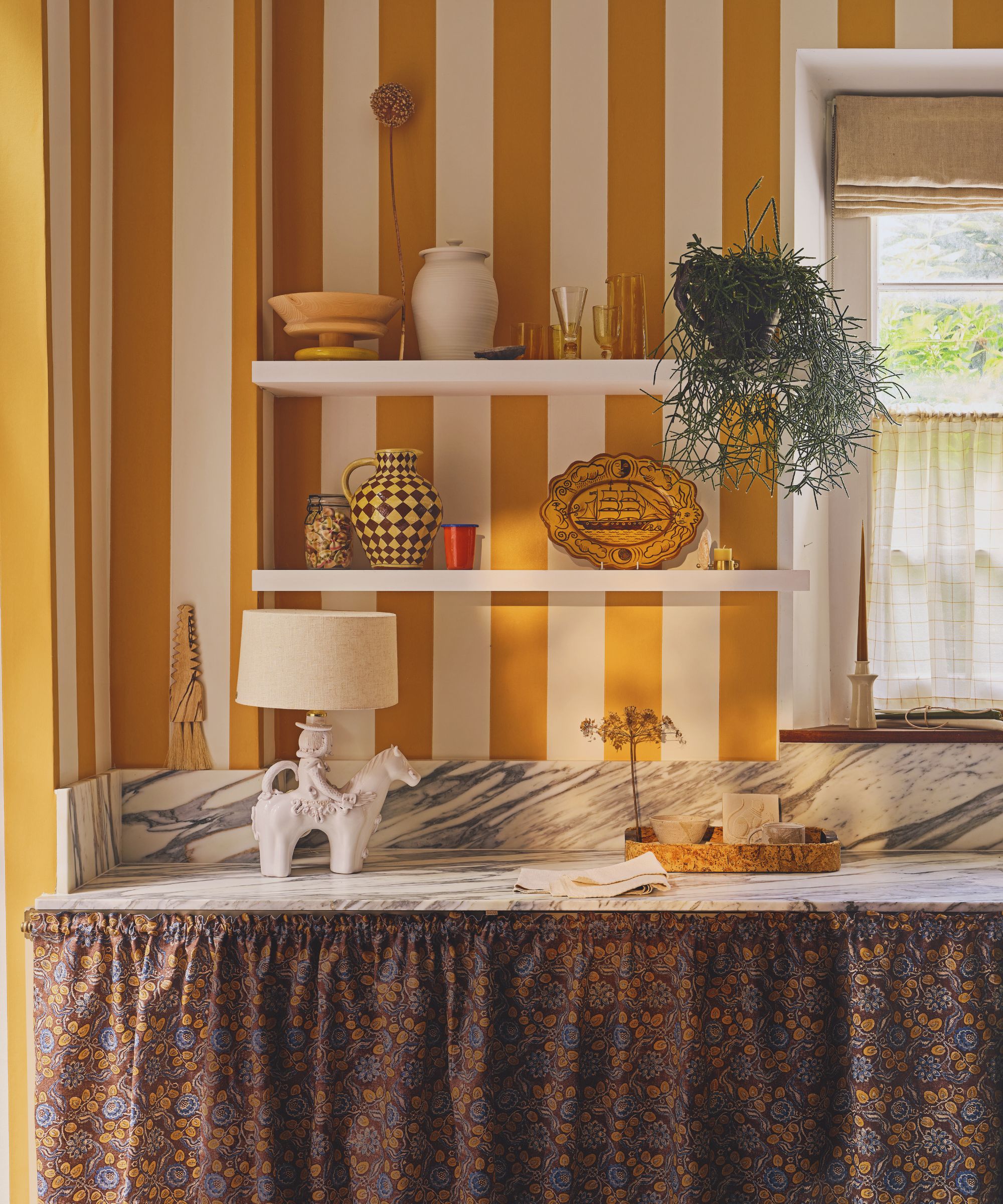 small kitchen with bold orange and white striped wallpaper, with marble countertops and an orange floral patterned cabinet skirt styled with a lamp and open shelves