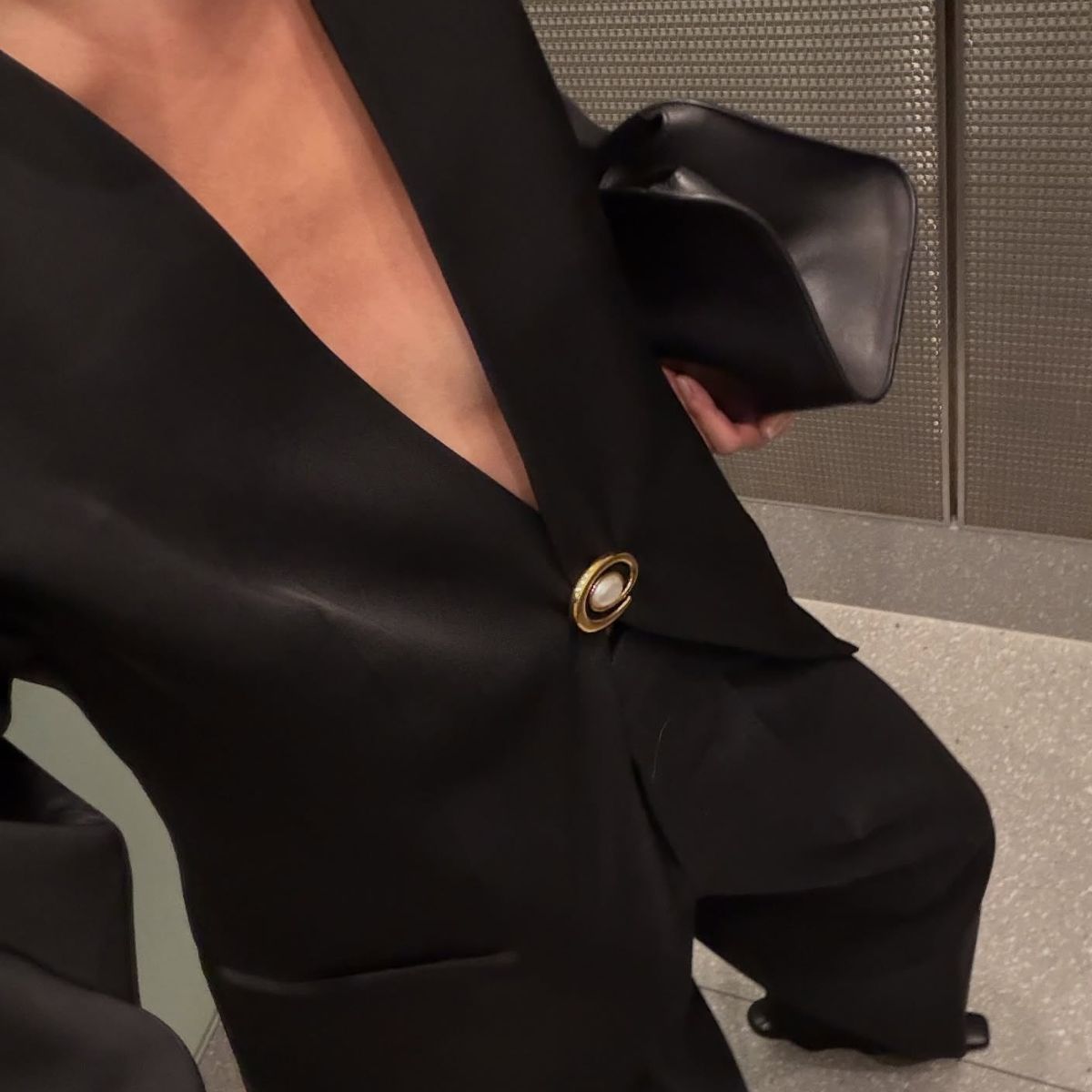 A woman taking a selfie of her outfit wearing a black blazer, black trousers, black heeled boots and an oversized black clutch bag