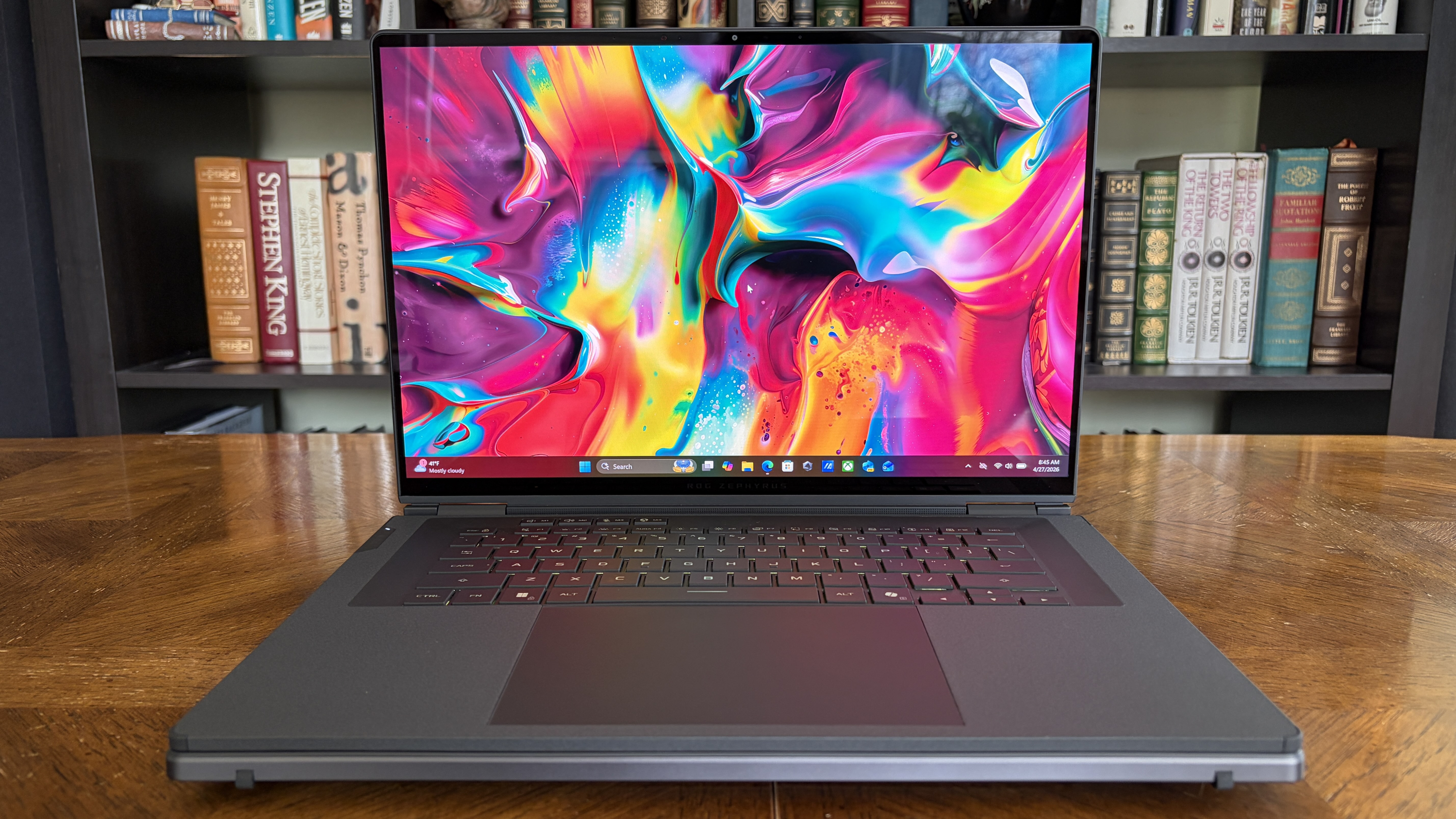 Laptop displaying vibrant, swirling abstract art in pink, blue, and yellow hues. It rests on a wooden table with bookshelves in the background.