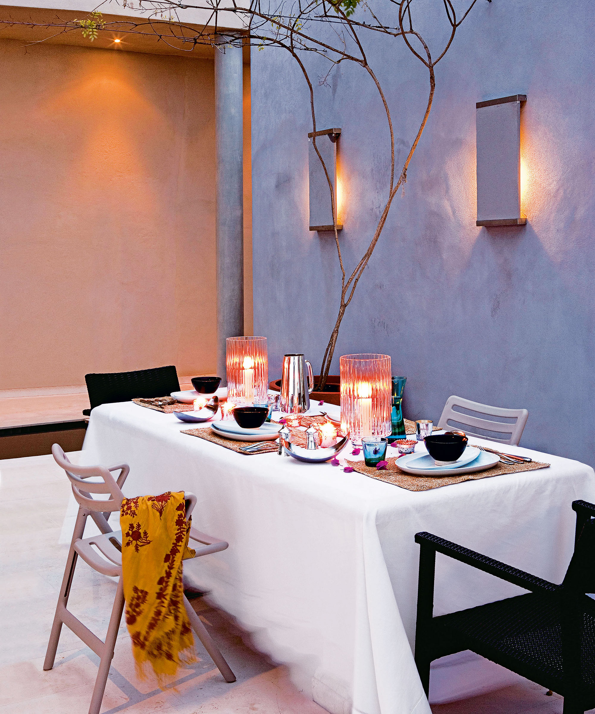 Wall lighting and table-top candles in a courtyard garden with whitewashed plaster walls and dining table and chairs.