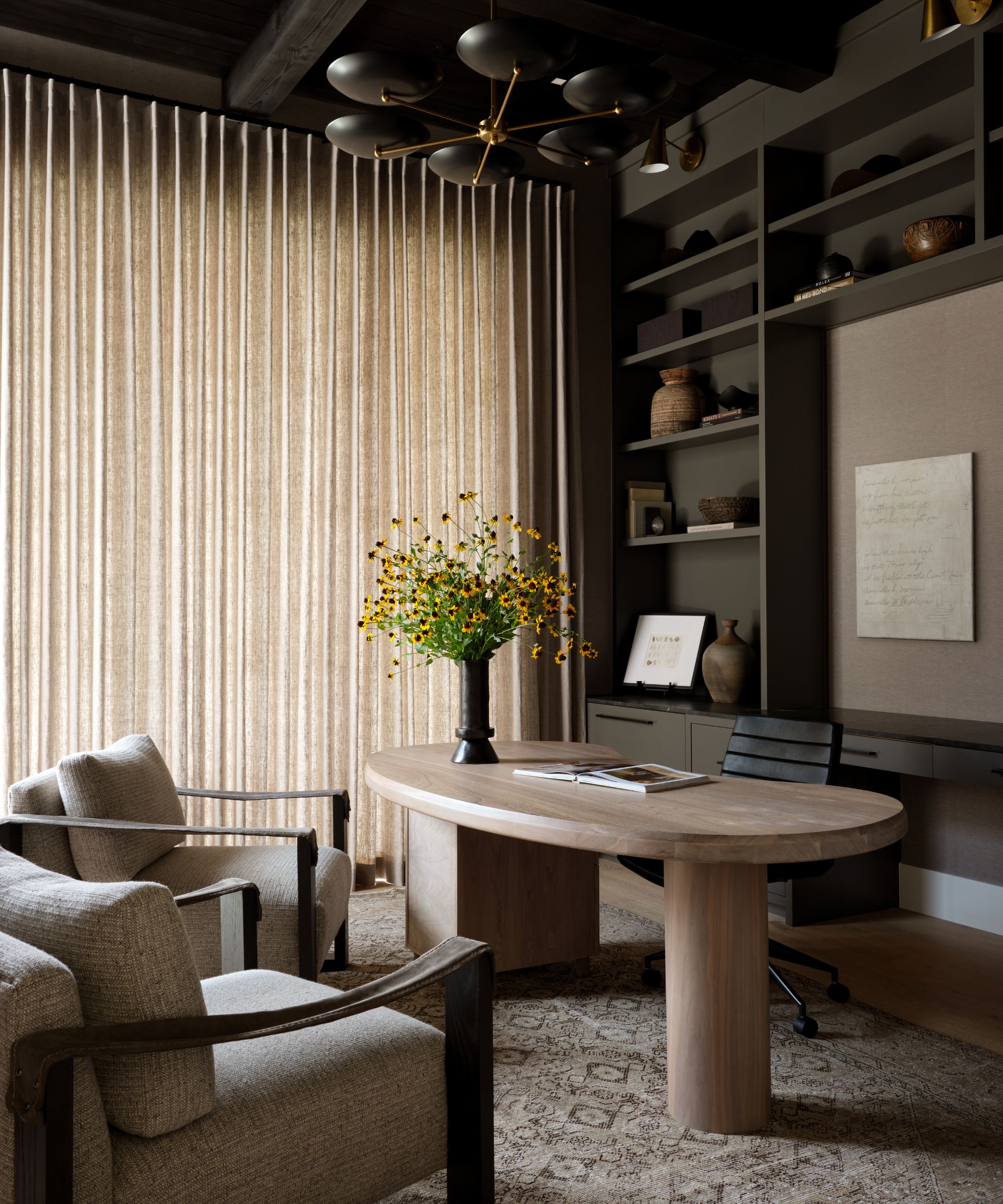 A home office color-drenched in dark brown paint with a bean-shaped natural wood desk and wall shelves displaying books and decor