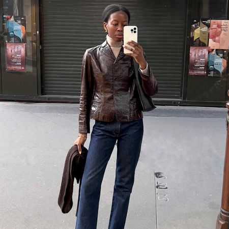 Sylvie Mus wears a brown leather short jacket with a beige turtleneck, blue jeans, and black shoulder bag.