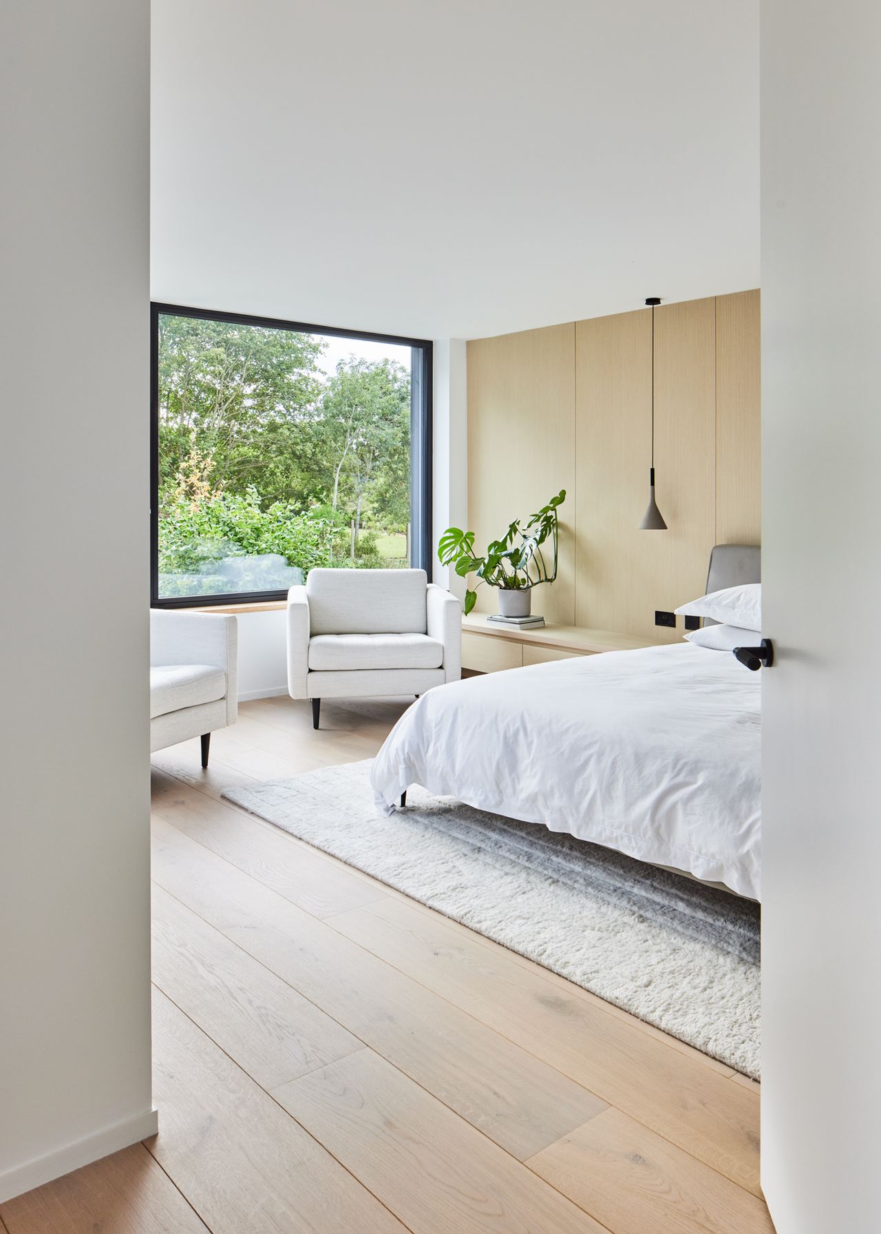 A contemporary bedroom with a double bed, plywood wall, and a large square window with views out to a garden beyond
