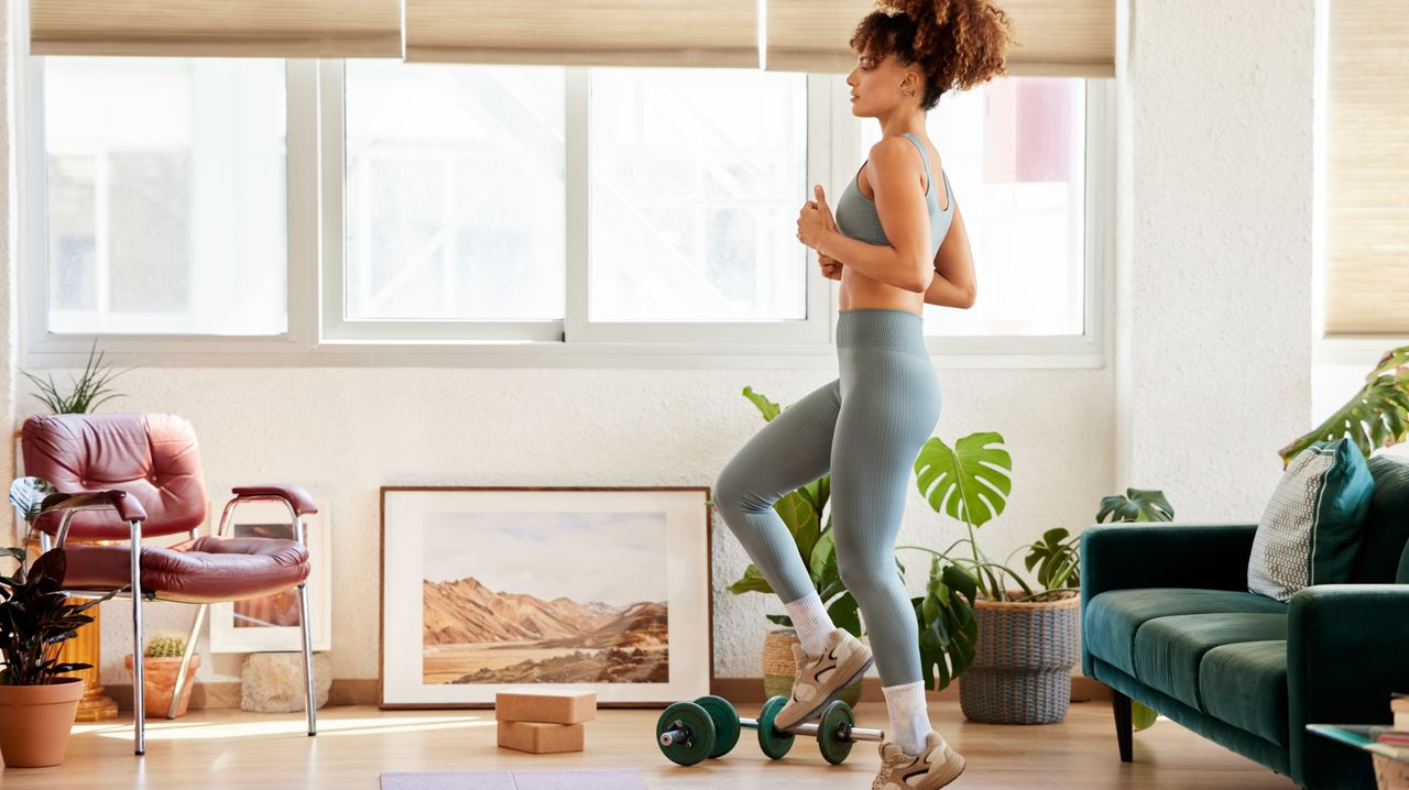 A woman does a walking workout in her living room. She is marching on the spot with her arms bent and held at her side. She wears grey leggings, a tank top and sneakers. Behind her we see plants, a couch, a chair, and paintings. There are also dumbbells and yoga blocks to her side.