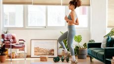 A woman does a walking workout in her living room. She is marching on the spot with her arms bent and held at her side. She wears grey leggings, a tank top and sneakers. Behind her we see plants, a couch, a chair, and paintings. There are also dumbbells and yoga blocks to her side.