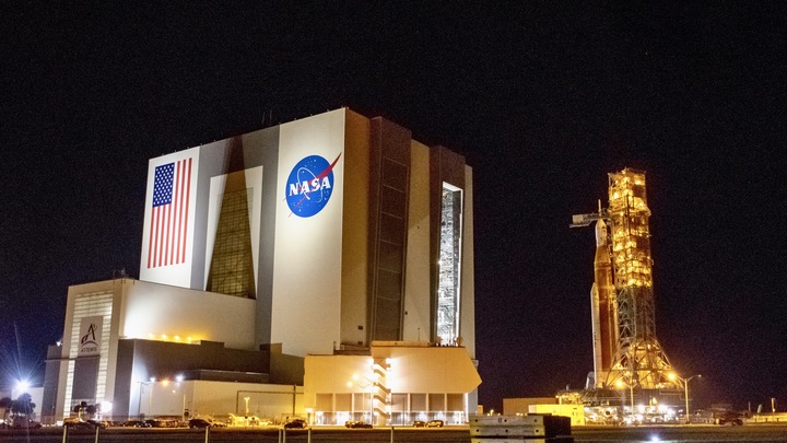 A rocket on a mobile launch platform stands outside a massive cube building at night.