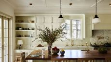 A bright, airy kitchen with cream-colored cabinetry, a large dark wood island with barstool seating, and marble countertops. Two white pendant lights hang above the island, and a large vase of greenery sits as a centerpiece near a window.