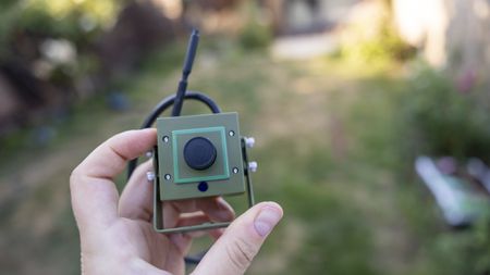 A female hand holding the Green Feathers HD WiFi bird box camera in a summer garden
